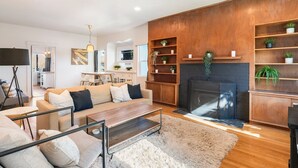 A modern living room with a sofa, coffee table, and fireplace. There are shelves with plants and a TV mounted on the wall.