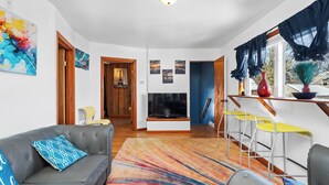 A modern living room with a grey sofa, colorful rug, and a TV. There are yellow and white chairs, a wooden door, and a blue wall. - Charming 3-bedroom duplex with study space, WiFi in cool  Scituate. (Scituate)