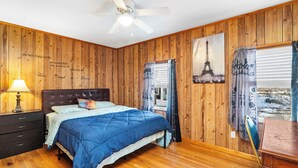 A bedroom with a wooden paneled wall, a bed with a blue comforter, a ceiling fan, and a window with curtains. - Charming 3-bedroom duplex with study space, WiFi in cool  Scituate. (Scituate)