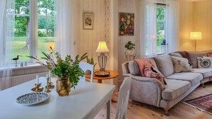 A cozy living room with a white table, a grey sofa, and a lamp. There are windows with curtains, a painting on the wall, and a plant on a side table. - 3 bedroom stunning home in Tingsryd (Tingsryd)