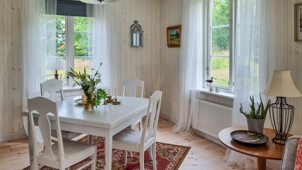 A dining area with a white table and chairs, a red patterned rug, and a view of greenery outside through large windows. - 3 bedroom stunning home in Tingsryd (Tingsryd)