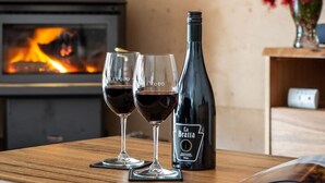 Una mesa de madera con dos copas de vino y una botella, una chimenea al fondo, y un jarrón con flores sobre una estantería.