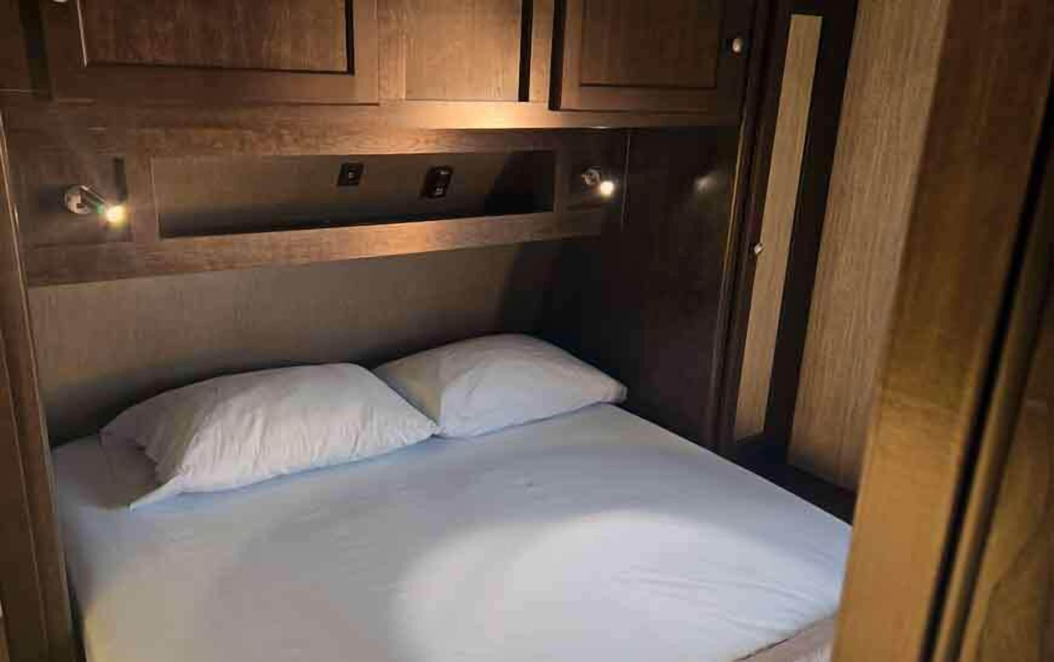 A compact bed with white bedding in a small, enclosed space with wooden walls and cabinets above.