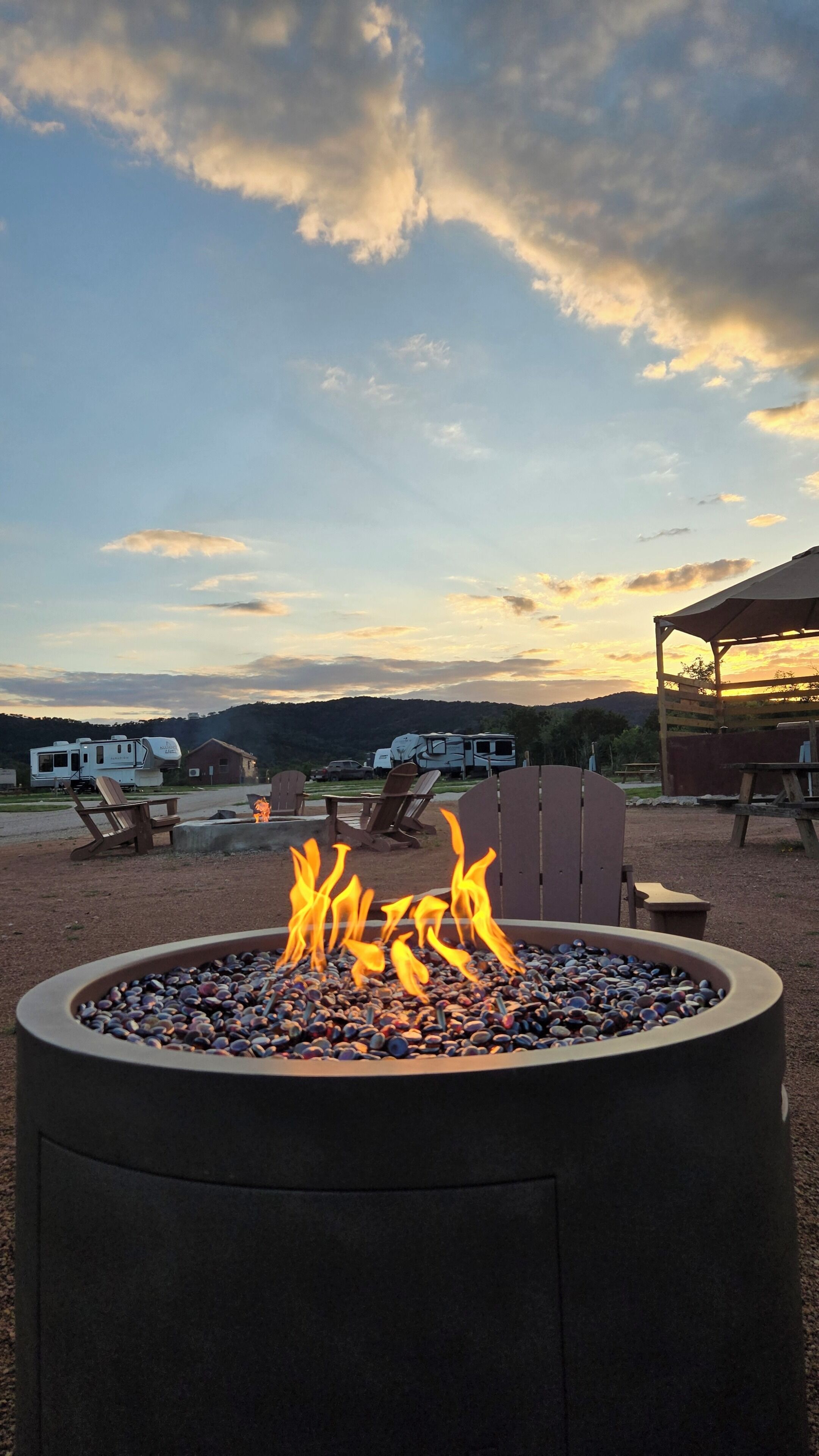 A fire pit with flames, surrounded by outdoor seating and RVs in a campground setting.