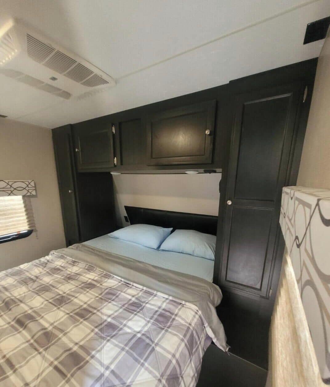 A bedroom with a bed, a headboard, a window with blinds, and a wall-mounted air vent.
