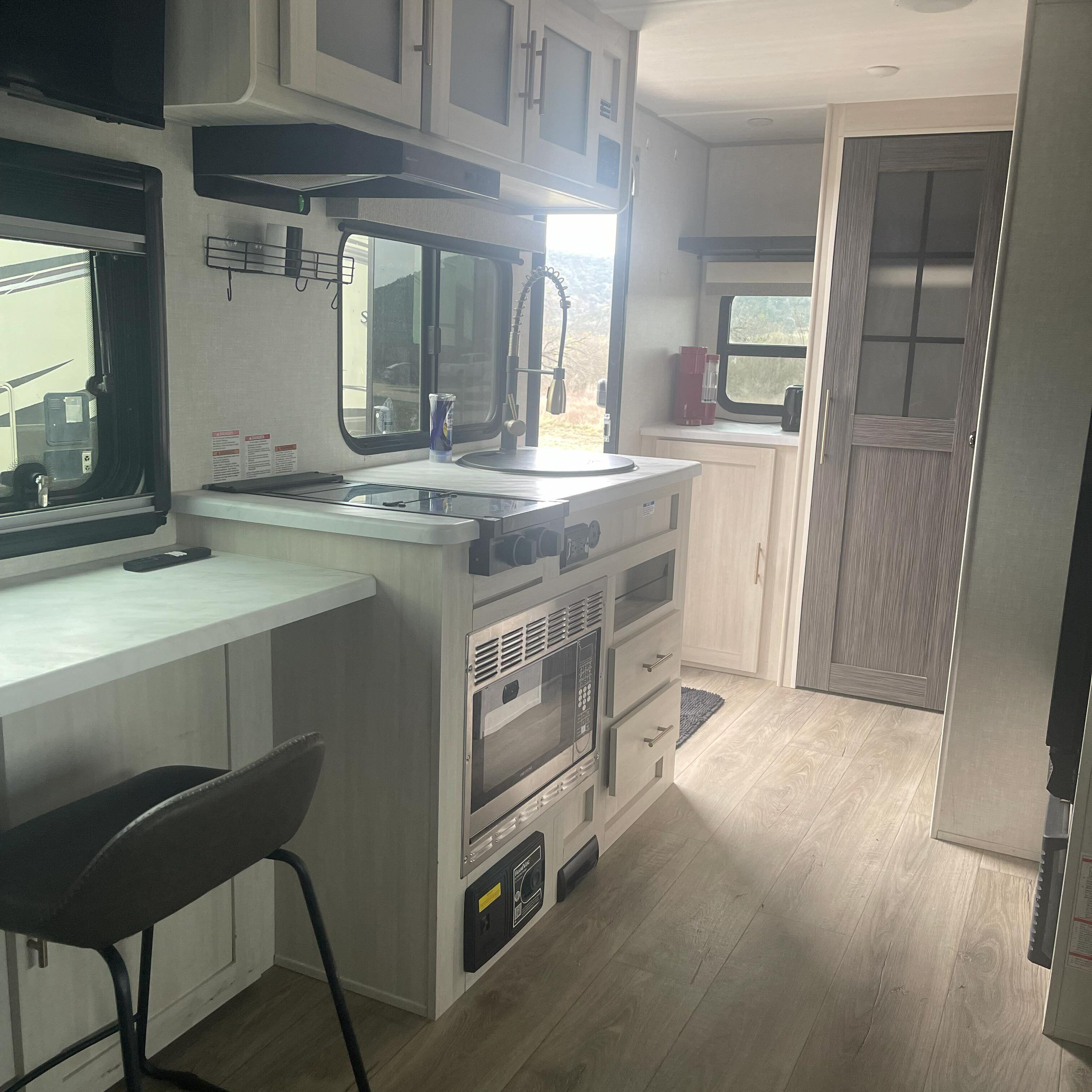 A compact kitchen with a built-in oven, microwave, and sink. There is a chair and a small table, and a window providing a view outside.