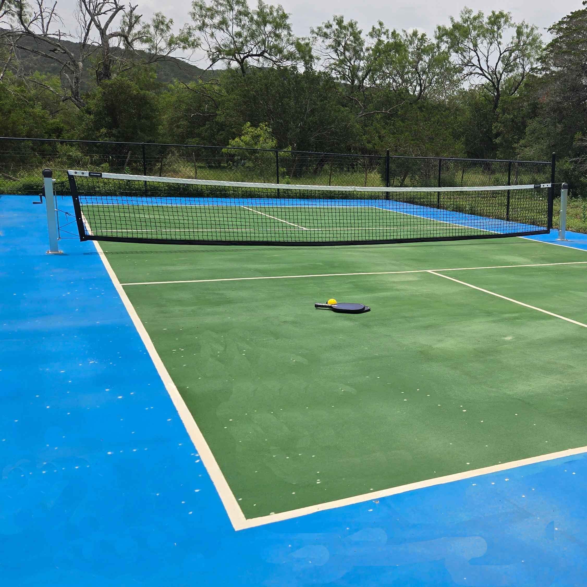 Cancha de tennis a l'aire lliure amb xarxa, envoltada d'arbres i superfície blava.