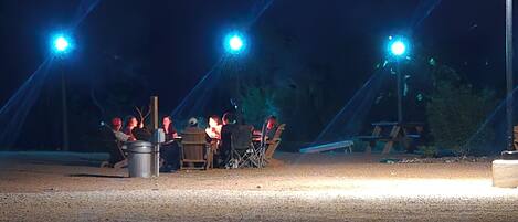 An outdoor dining area with tables and chairs, illuminated by overhead lights.