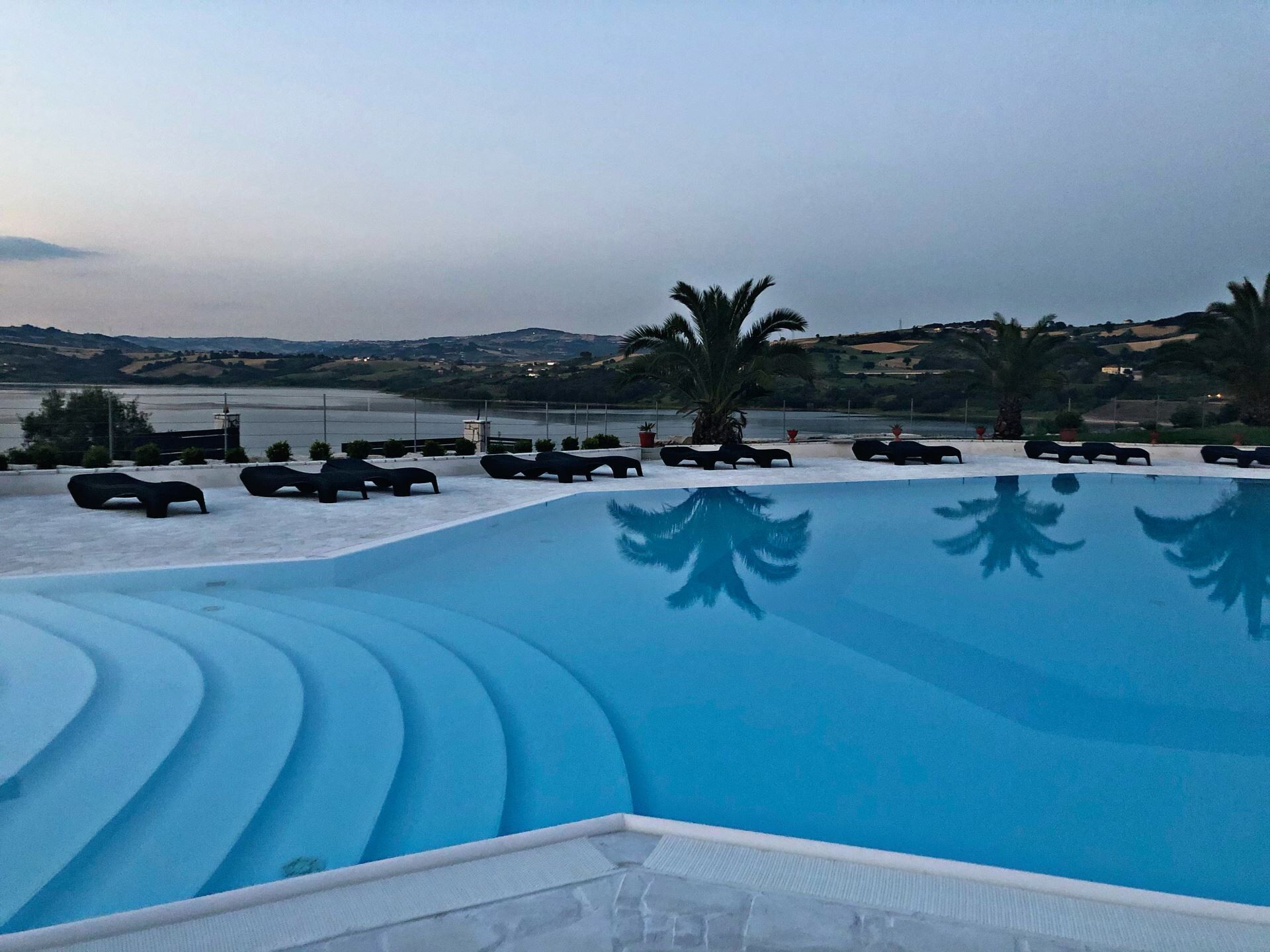 Ein Pool mit Liegestühlen und Palmen, umgeben von hügeliger Landschaft und einem Gewässer.
