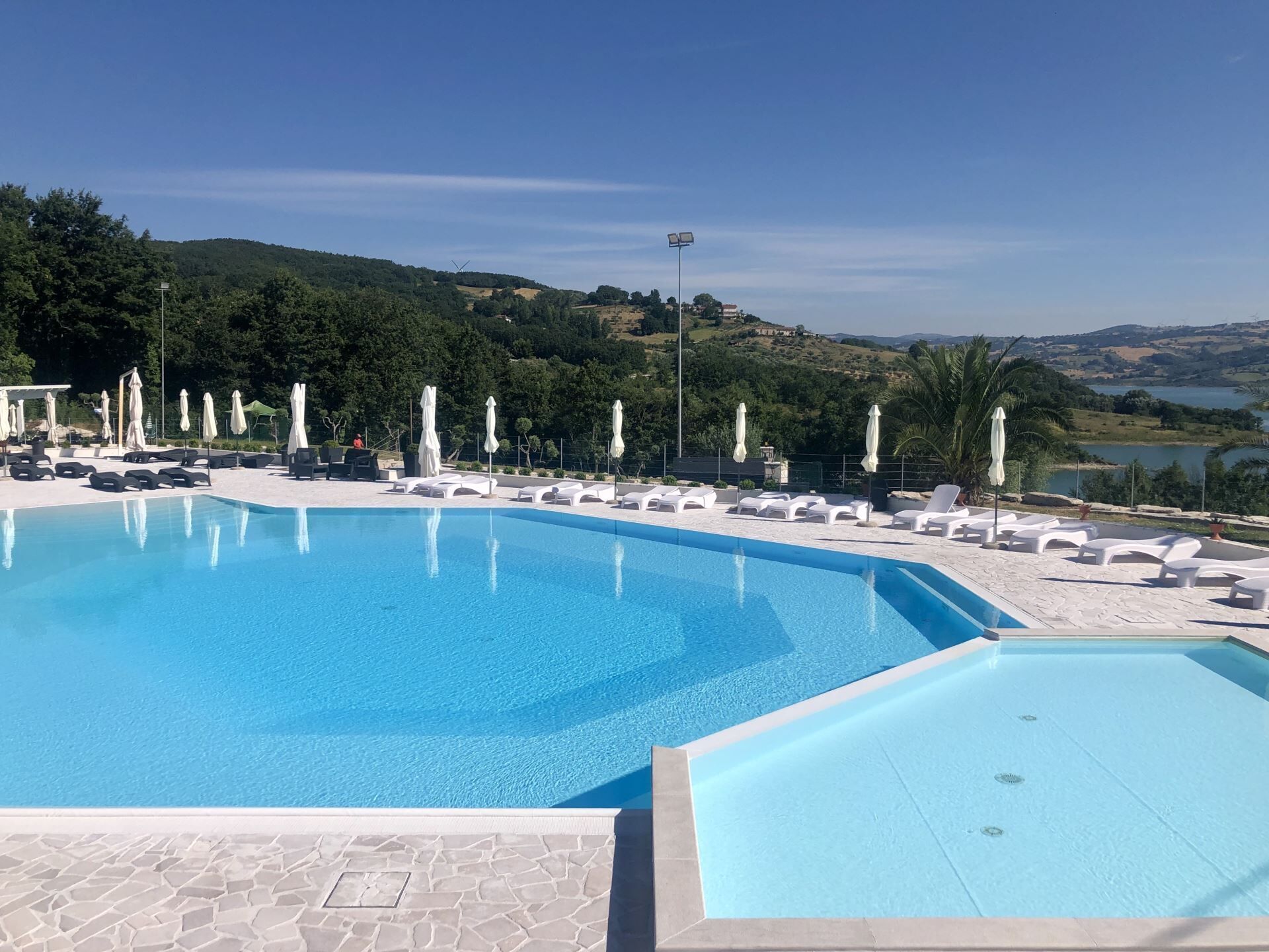 A swimming pool with lounge chairs and umbrellas, surrounded by a landscaped area with a view of hills and a body of water.