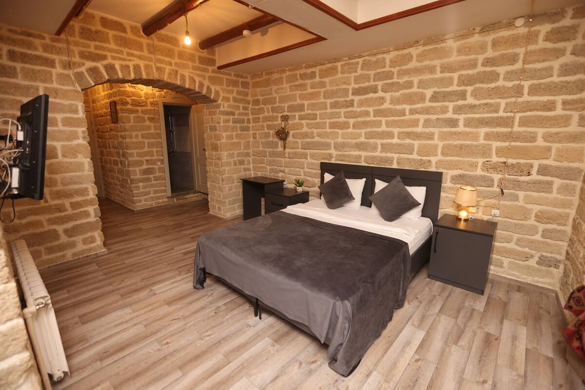 Ein Schlafzimmer mit einer Steinsäule, einem Bett mit grauer Bettdecke, einem Nachttisch mit Lampe und einer kleinen Pflanze.