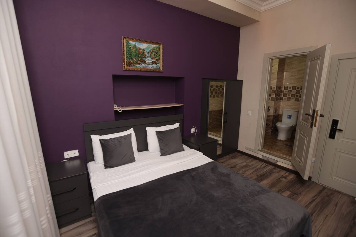 Ein Schlafzimmer mit Bett, Nachttisch, Kommode, einem Bild an der Wand und einer Badezimmertür.