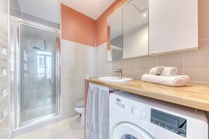 A modern bathroom with a glass shower enclosure, a white toilet, a sink with a wooden countertop, and a washing machine. - Appartement Antonin - Welkeys (Villeurbanne)