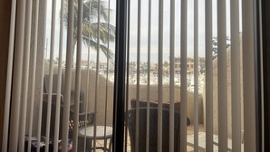 Een kamer met uitzicht door verticale zonwering, met een balkon met buitenmeubilair en een verafgelegen uitzicht op gebouwen en palmbomen.