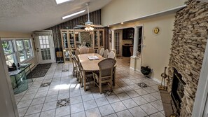 Een eetruimte met een grote tafel en stoelen, een stenen open haard, en uitzicht op de omgeving via een deur.