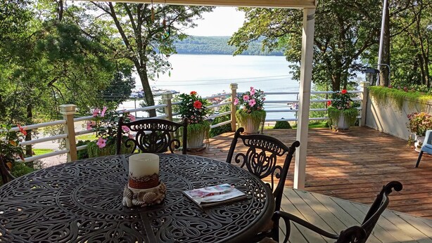 A covered outdoor seating area with a view of a lake, surrounded by greenery and a white railing. - Grandview Getaway with a Million Dollar View! (Lakeville)