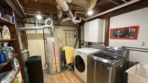 A laundry room with a washing machine, dryer, and water heater. There are pipes on the ceiling and a shelf with cleaning supplies. - Enjoy beautiful sunsets at a Lake Side Cottage (Tipton)