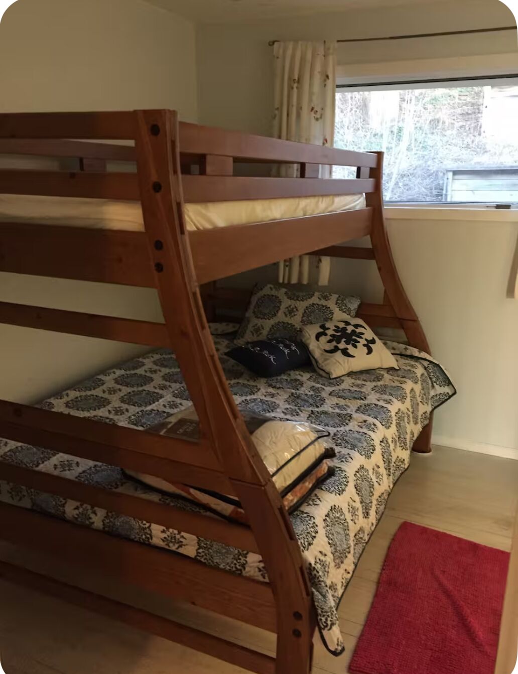 Een stapelbed met beddengoed en kussens met een patroon, een rood tapijt en een raam met gordijnen.
