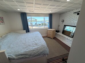A bedroom with a bed, a small dresser, a TV mounted on a brick wall, and a window with a view of the outdoors.