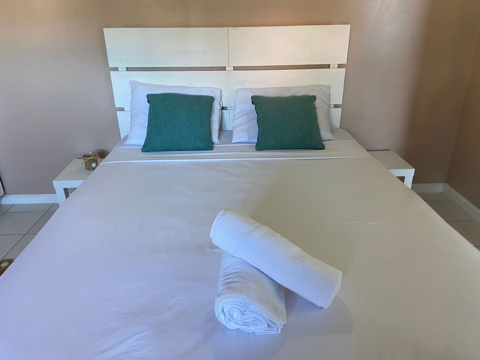 A neatly made bed with white linens and two green pillows, a white headboard, and a small bedside table with a lamp.