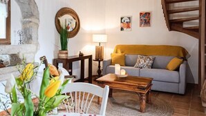 Un salon chaleureux avec des poutres apparentes, une cheminée en pierre, un canapé et une table à manger recouverte d’une nappe à motifs floraux.