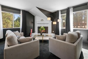 A modern living room with a fireplace, two armchairs, a coffee table, and large windows with curtains.