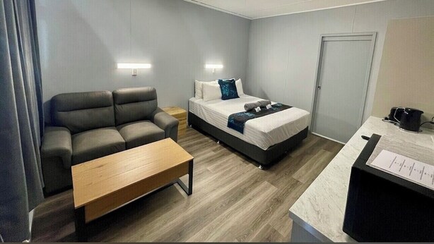 Ein modernes Hotelzimmer mit grauem Sofa, einem Holztisch, einem Bett mit weißer Matratze und blauen Kissen, einem Nachttisch und einem Schreibtisch mit Computer und Papieren.