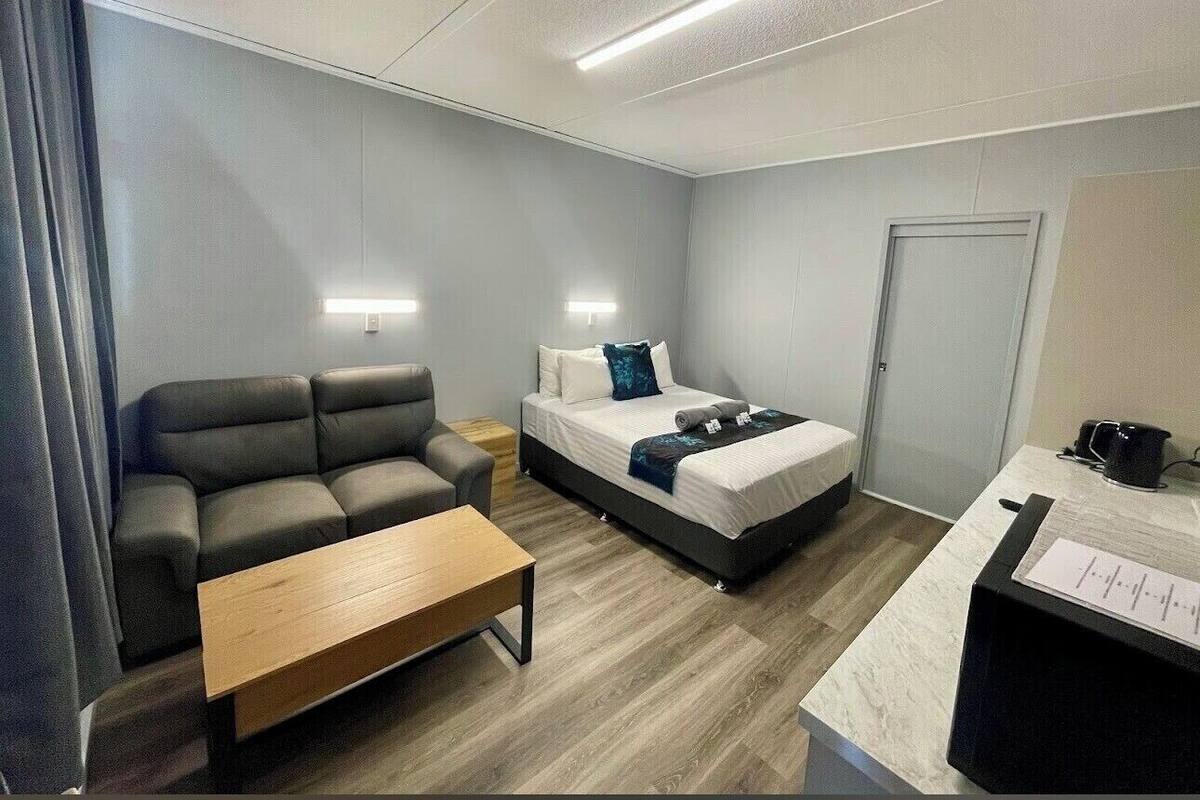 Una habitación de hotel moderna con un sofá gris, una mesa de centro de madera, una cama con colchón blanco y almohadas azules, una mesita de noche y un escritorio con computadora y documentos.