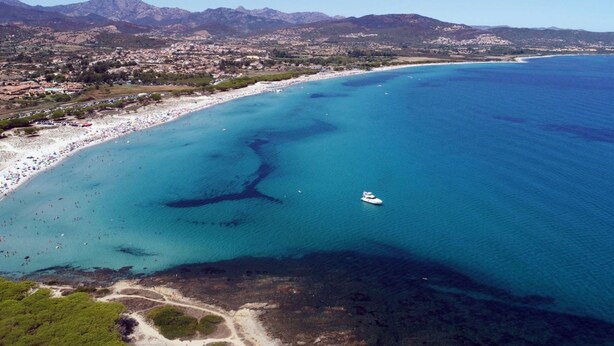 An aerial view of a coastal area with a beach, clear blue waters, and a mountainous backdrop.