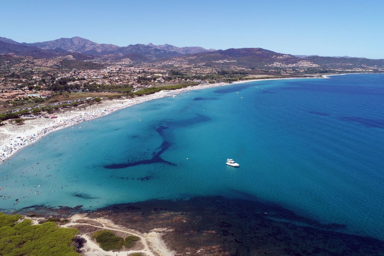 Vista aérea de una zona costera con playa, aguas azules cristalinas y montañas al fondo.