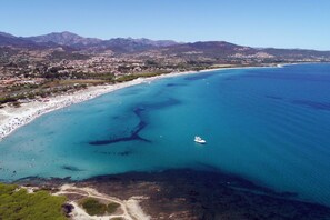 An aerial view of a coastal area with a beach, clear blue waters, and a mountainous backdrop.