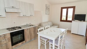 A compact kitchen with a gas stove, oven, and sink. There is a dining table with chairs and a sofa in the adjacent living area. A TV is mounted on the wall.