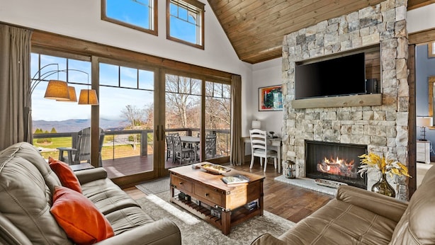 A spacious living room with a stone fireplace, leather sofas, and a large window overlooking a scenic view. - Sommer Hus at Eagles Nest: Pet Friendly Single Level (Banner Elk)