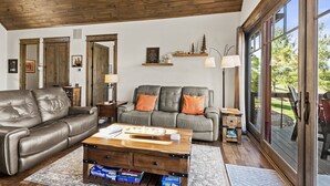 A spacious living room with a leather sofa, a wooden coffee table, and a grey couch with orange cushions. There are wooden shelves with decorative items and a large window providing a view of the outdoors. - Sommer Hus at Eagles Nest: Pet Friendly Single Level (Banner Elk)