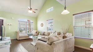 A living room with a beige sofa, a glass coffee table, and a ceiling fan. There are two windows with blinds, a small rug, and a wall-mounted clock. - Comfortable Emerald Coast Retreat! Only 3 minute walk to the white sandy BEACH! (Panama City Beach)