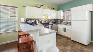 A kitchen with white cabinets, a countertop island, wooden stools, a surfboard decoration, and a refrigerator. - Comfortable Emerald Coast Retreat! Only 3 minute walk to the white sandy BEACH! (Panama City Beach)