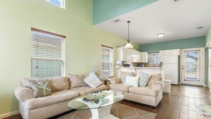 A living room with a beige sofa, a glass coffee table, and a kitchen in the background. - Comfortable Emerald Coast Retreat! Only 3 minute walk to the white sandy BEACH! (Panama City Beach)