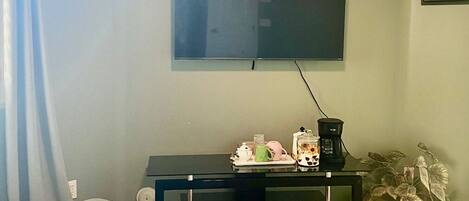 A living room with a flat-screen TV mounted on the wall, a glass-top coffee table, a dark entertainment unit, and a potted plant.