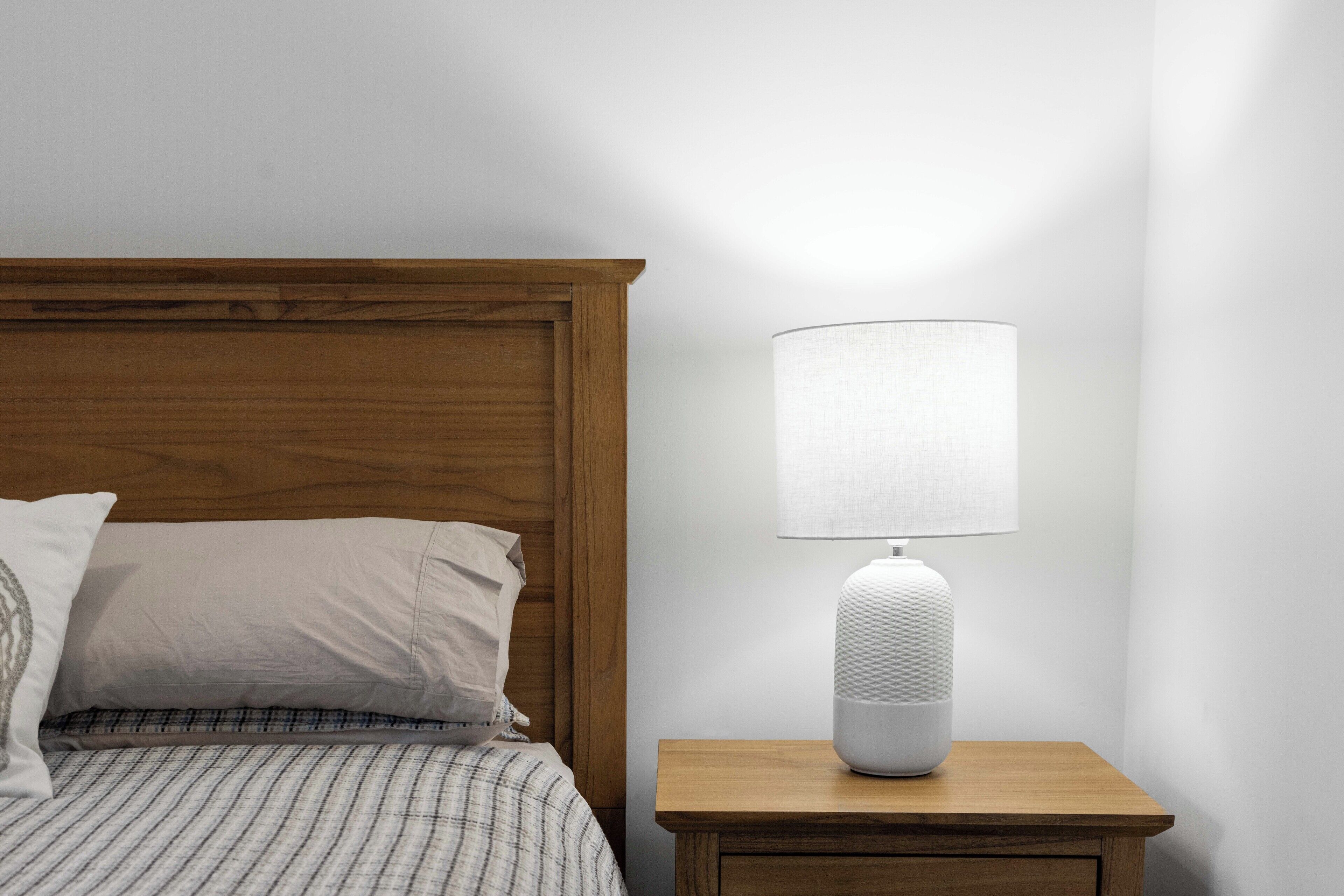 A neatly made bed with a wooden headboard, a bedside table with a white lamp, and a plain wall.