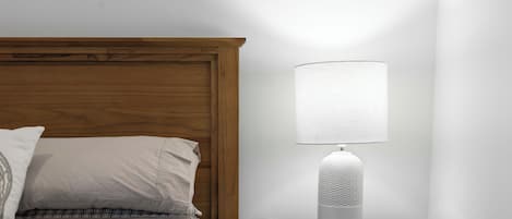 A neatly made bed with a wooden headboard, a bedside table with a white lamp, and a plain wall.