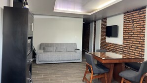 A modern living room with a grey sofa, a wooden dining table with two chairs, a flat-screen TV mounted on a brick-patterned wall, and a black cabinet with a television on top. - Hermosa Province Duplex House (Large Groups) (Guadalajara)