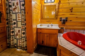 A wooden cabin bathroom with a sink, mirror, and red bathtub. - Cozy Cabin w Game Room hot tub Creek (Gatlinburg)