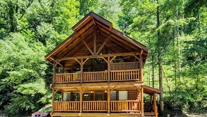 A two-story wooden cabin with a balcony, surrounded by dense forest.