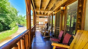 A wooden porch with a railing, outdoor seating, and a view of a road and greenery.