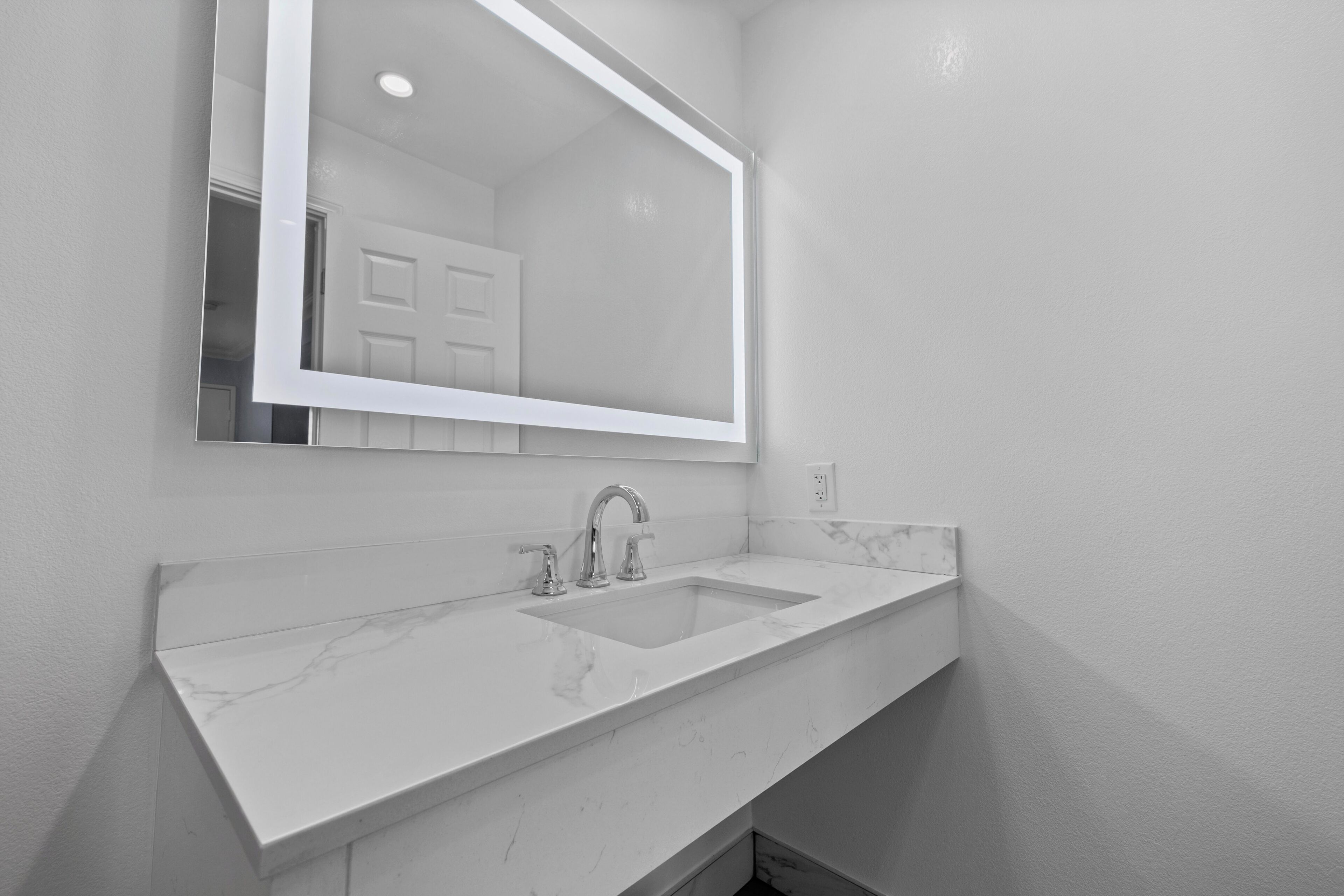 Un baño con lavamanos de mármol blanco, un espejo grande y una puerta al fondo.