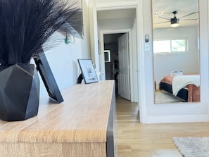 A room with a wooden desk, a black vase, and a framed picture. There is a bed with a white blanket and a pink pillow in the background.