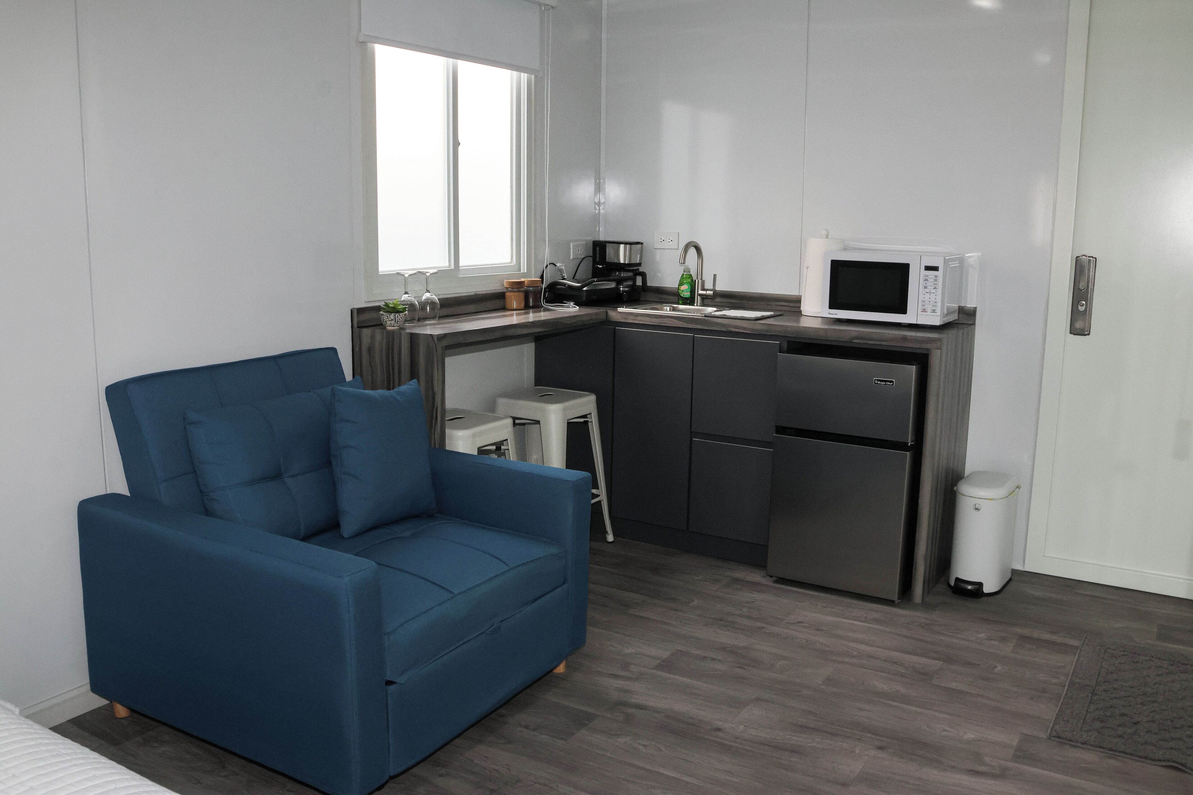 A compact kitchen with a sofa, a coffee machine, a microwave, and a trash bin.