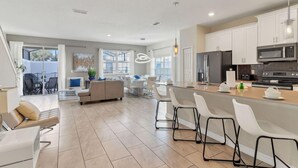 A modern kitchen with white cabinets, a central island with bar stools, and a dining area with a sofa and chairs. - Your 5bdr House w Pool - 4959 (Kissimmee)