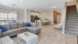 A modern living room with a sectional sofa, a white ottoman, and a dining area with a kitchen in the background. - Your 5bdr House w Pool - 4959 (Kissimmee)