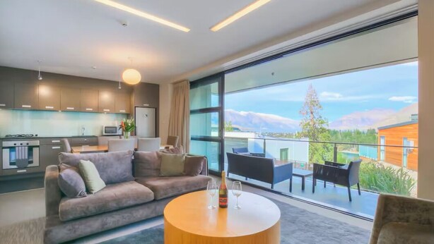 Una sala moderna con un sofá, una mesa de centro redonda de madera y un balcón con vistas a las montañas.
